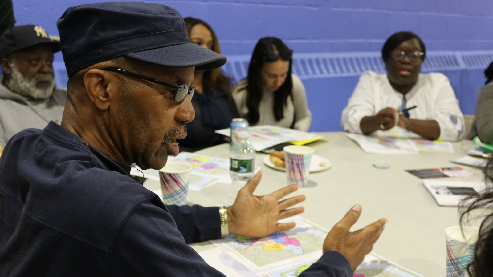 People sit around a white table covered in papers and drinks. One man in a hat and glasses is in the foreground.