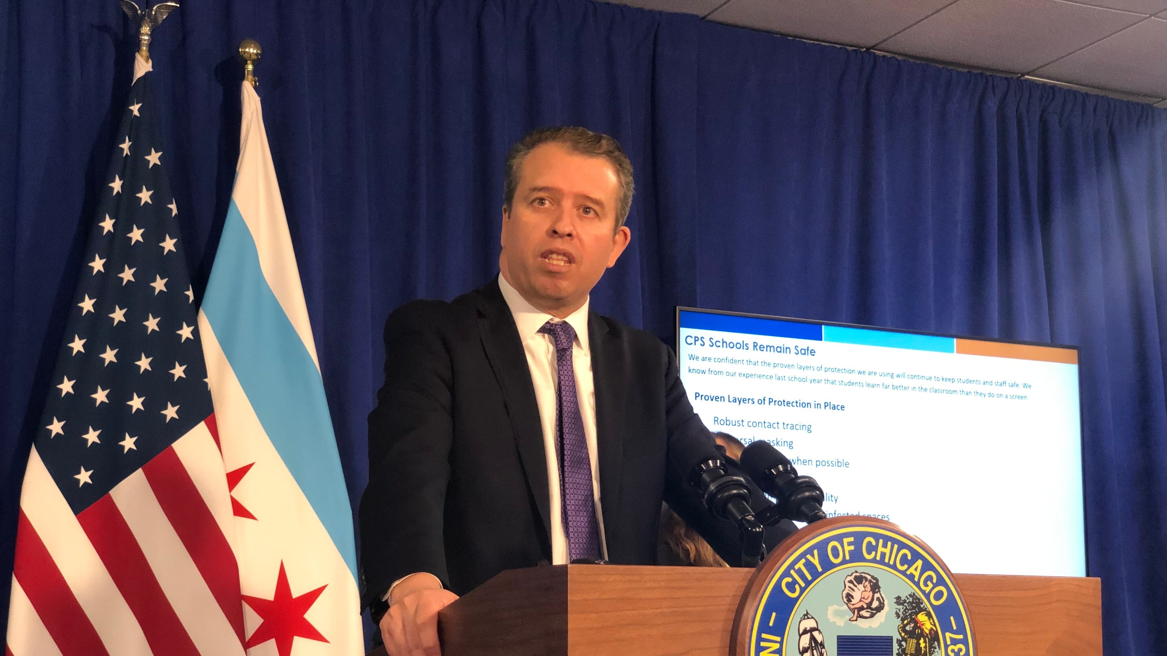 Chicago Public Schools CEO Pedro Martinez, wearing a black suit and purple tie, speaks at a City of Chicago podium during a press conference. Chicago and U.S. flags hang behind him against a blue backdrop, along with a screen providing COVID information.