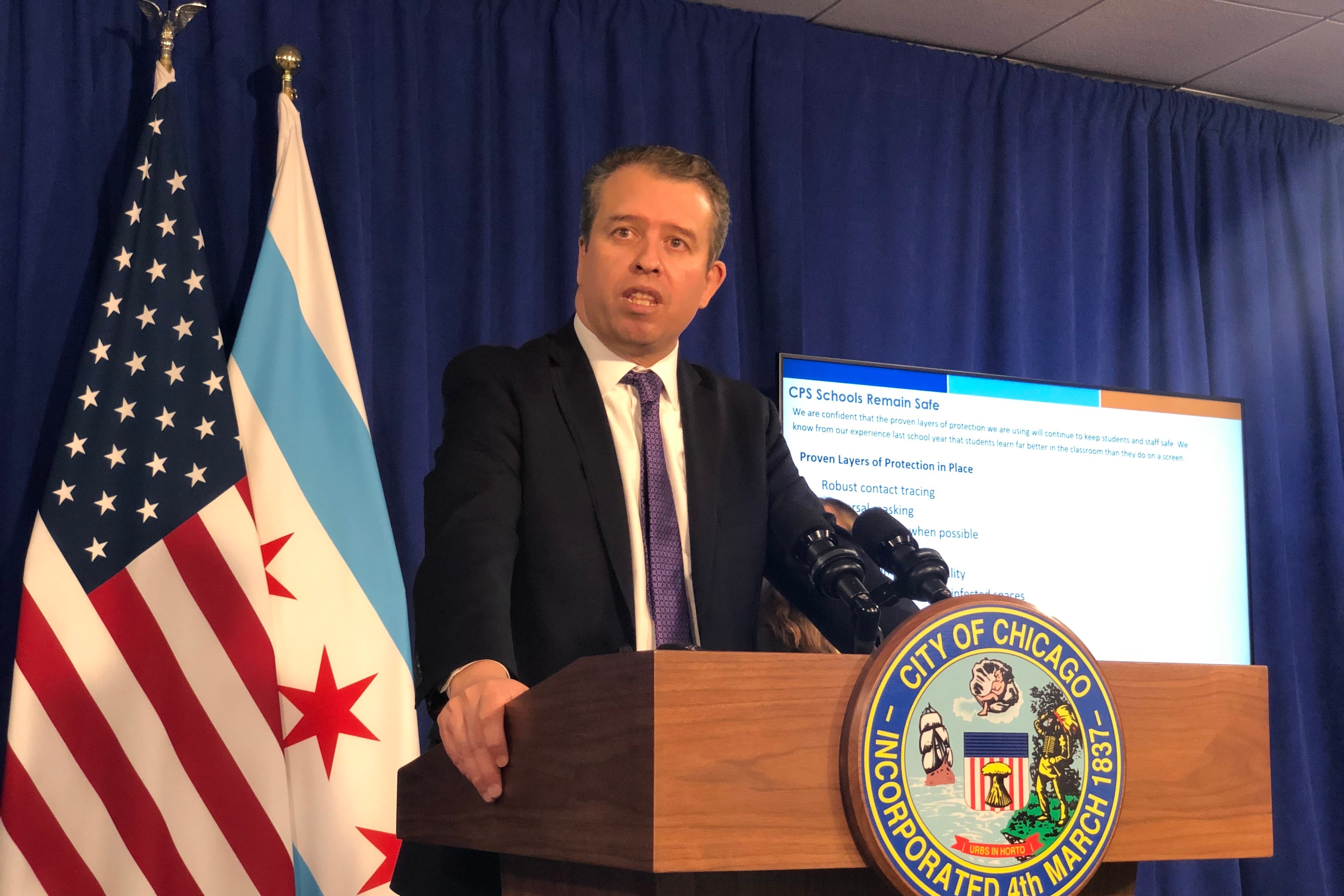 Chicago Public Schools CEO Pedro Martinez, wearing a black suit and purple tie, speaks at a City of Chicago podium during a press conference. Chicago and U.S. flags hang behind him against a blue backdrop, along with a screen providing COVID information.