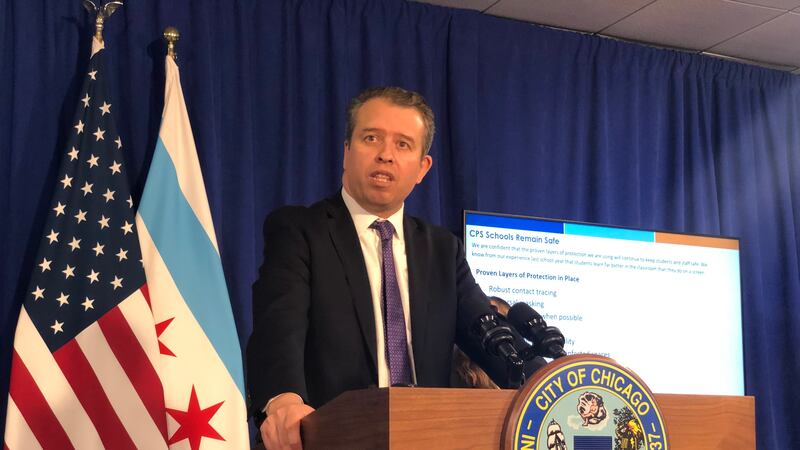 Chicago Public Schools CEO Pedro Martinez, wearing a black suit and purple tie, speaks at a City of Chicago podium during a press conference. Chicago and U.S. flags hang behind him against a blue backdrop, along with a screen providing COVID information.
