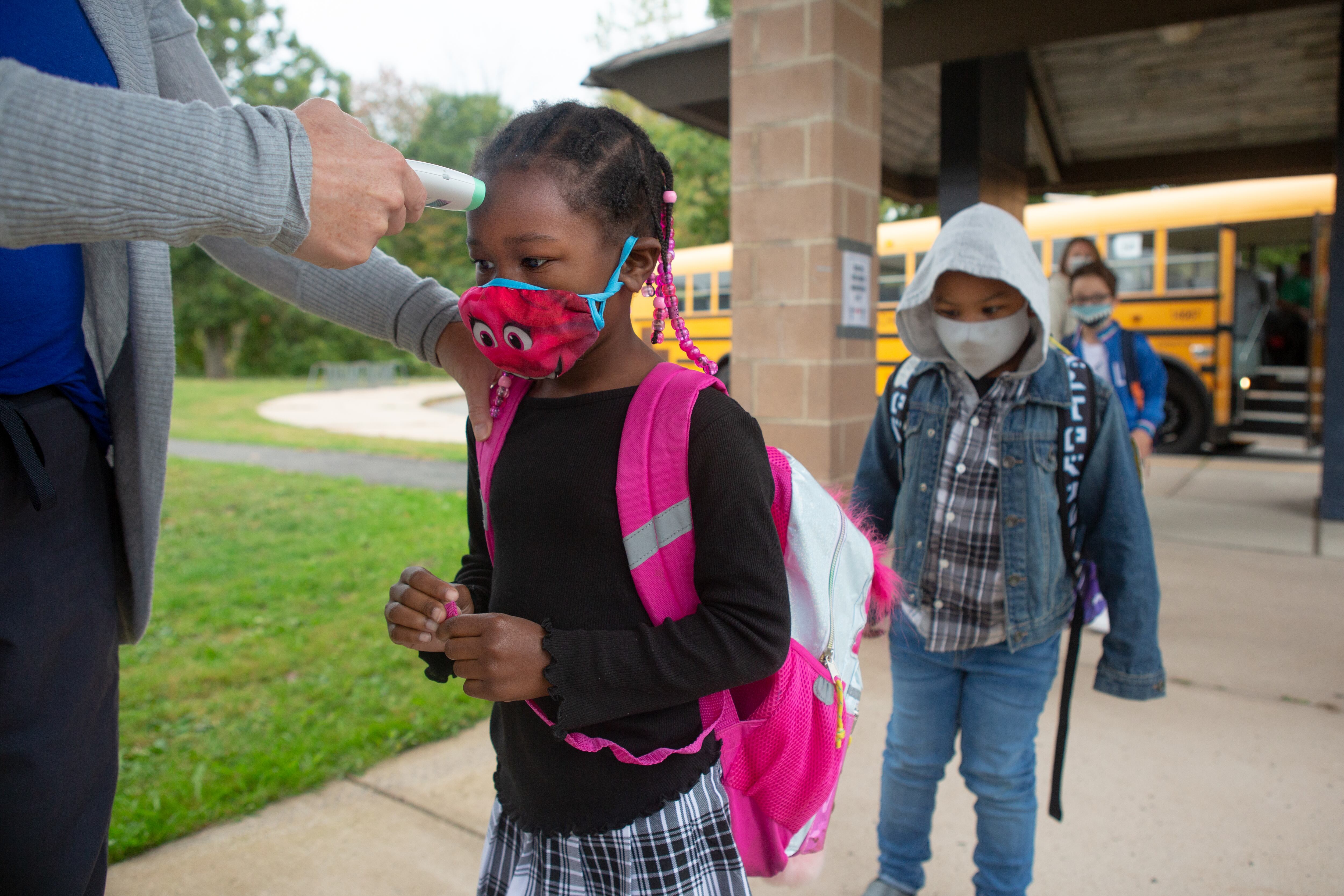 With a school bus in the background, students wearing masks and walking on a walkway receive temperature checks