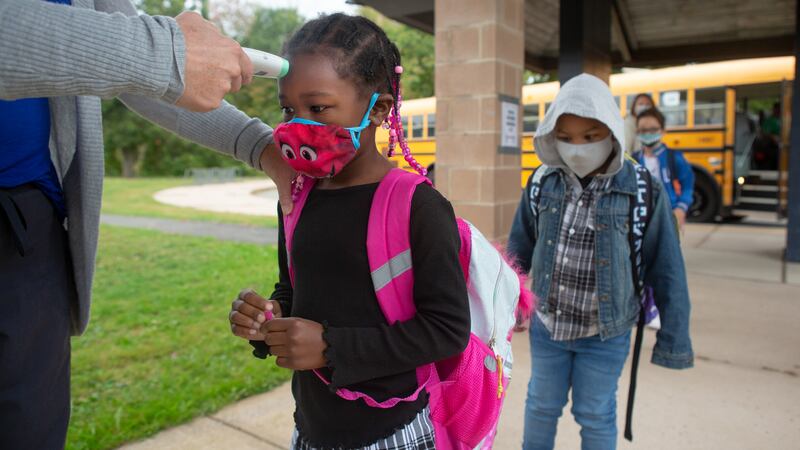 With a school bus in the background, students wearing masks and walking on a walkway receive temperature checks 