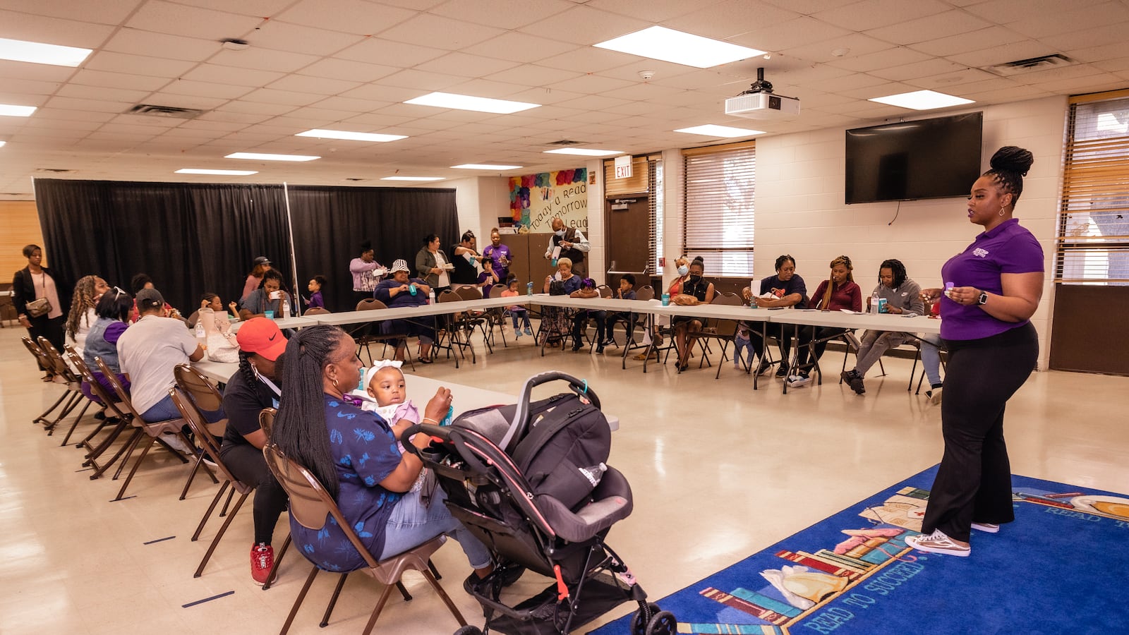 An educator wearing a purple shirt stands in front of a classroom. Sitting at tables around the room are a group of parents.