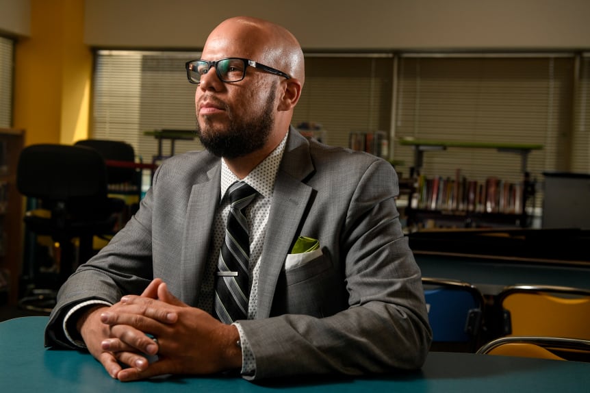 A portrait of Denver schools Superintendent Alex Marrero. He is sitting at a table with his hands crossed in front of him. He’s looking off to the side. He’s wearing glasses and a gray suit.