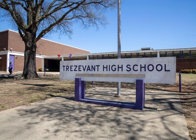 A large sign with the words "Trezevant High School" sit on a concrete slab in front of a large brick school building with a tree on the left and a blue sky in the background.
