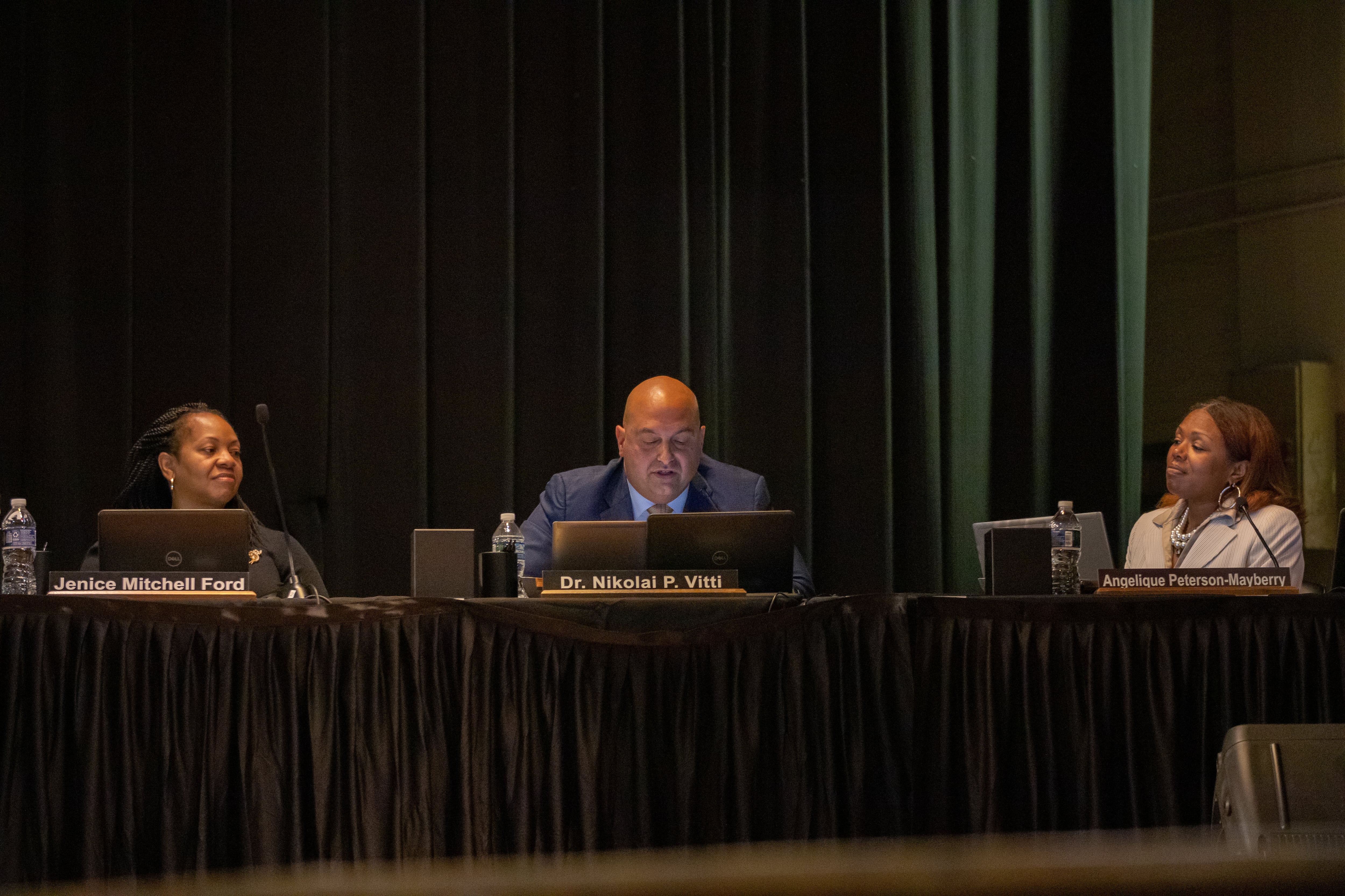 A man in a suit speaks on a school auditorium stage as two other people look at him.