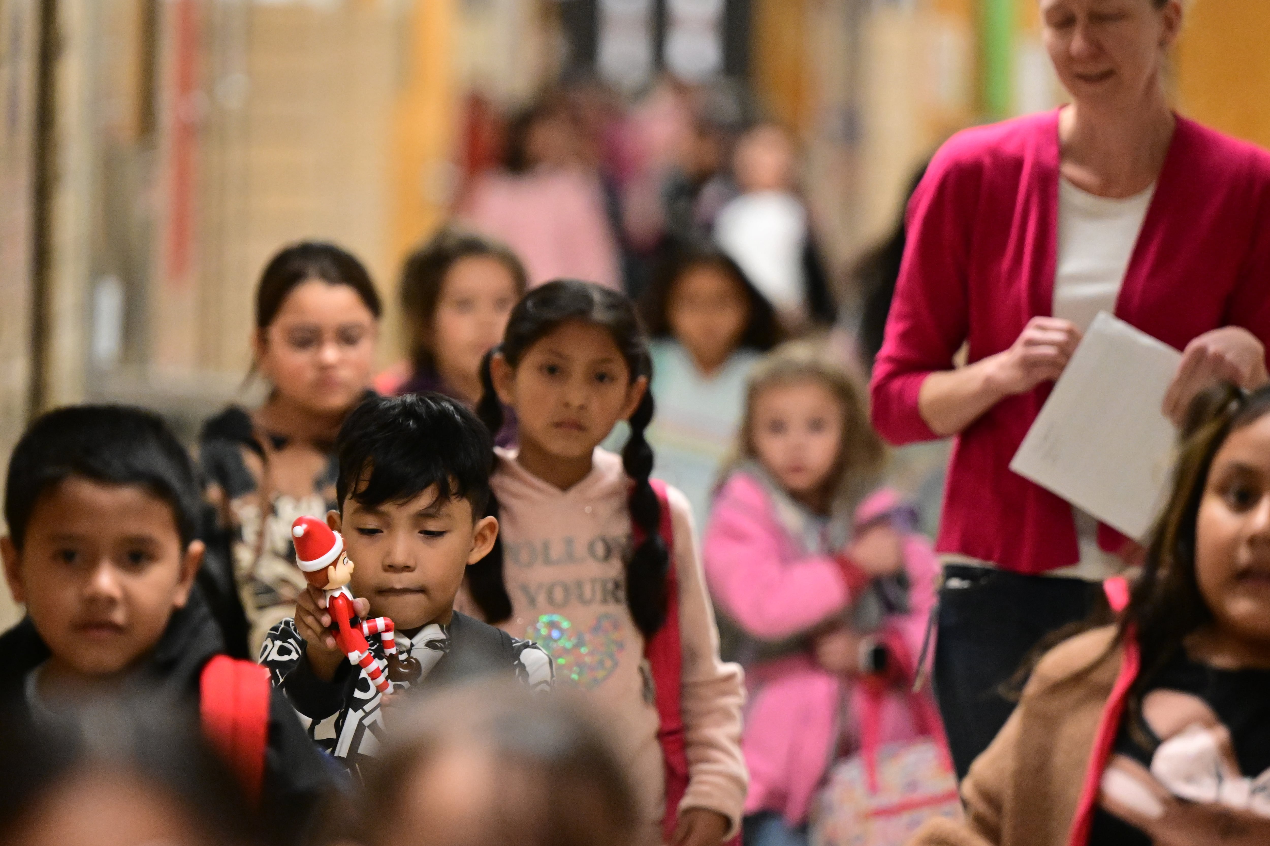 Young students move through a crowded school hallway. A girl near the center of the frame looks at the camera. She has large dark eyes and long dark braids.