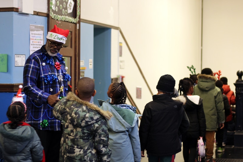 A man wearing a festive shirt and a Santa hat, talks with students as they walk by in a hallway.