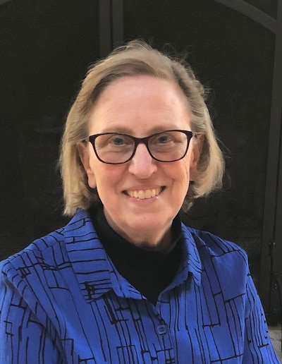 An older woman wearing a blue shirt and glasses smiles for a portrait.