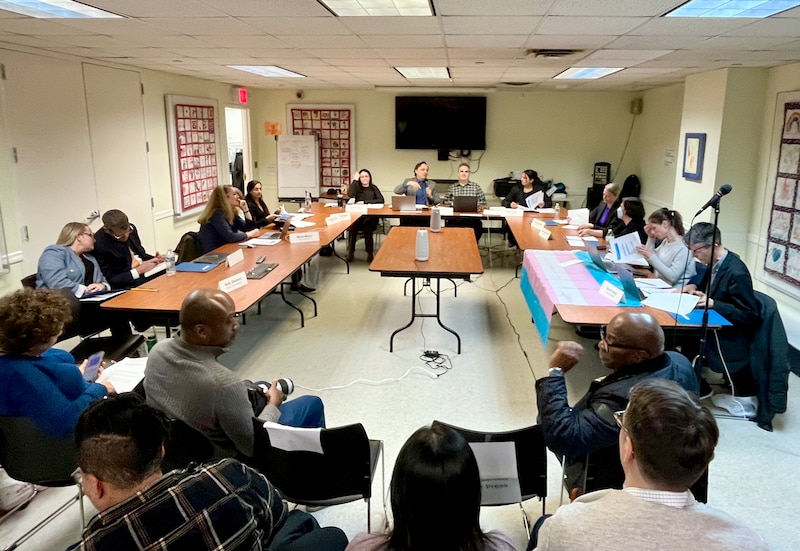 A group of people sit at tables in a rectangle in a room.