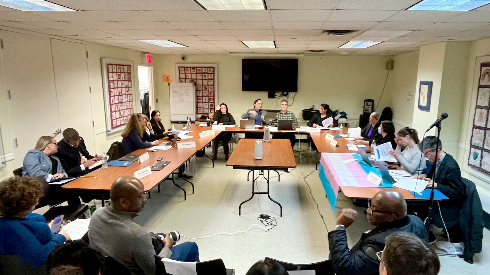 A group of people sit at tables in a rectangle in a room.
