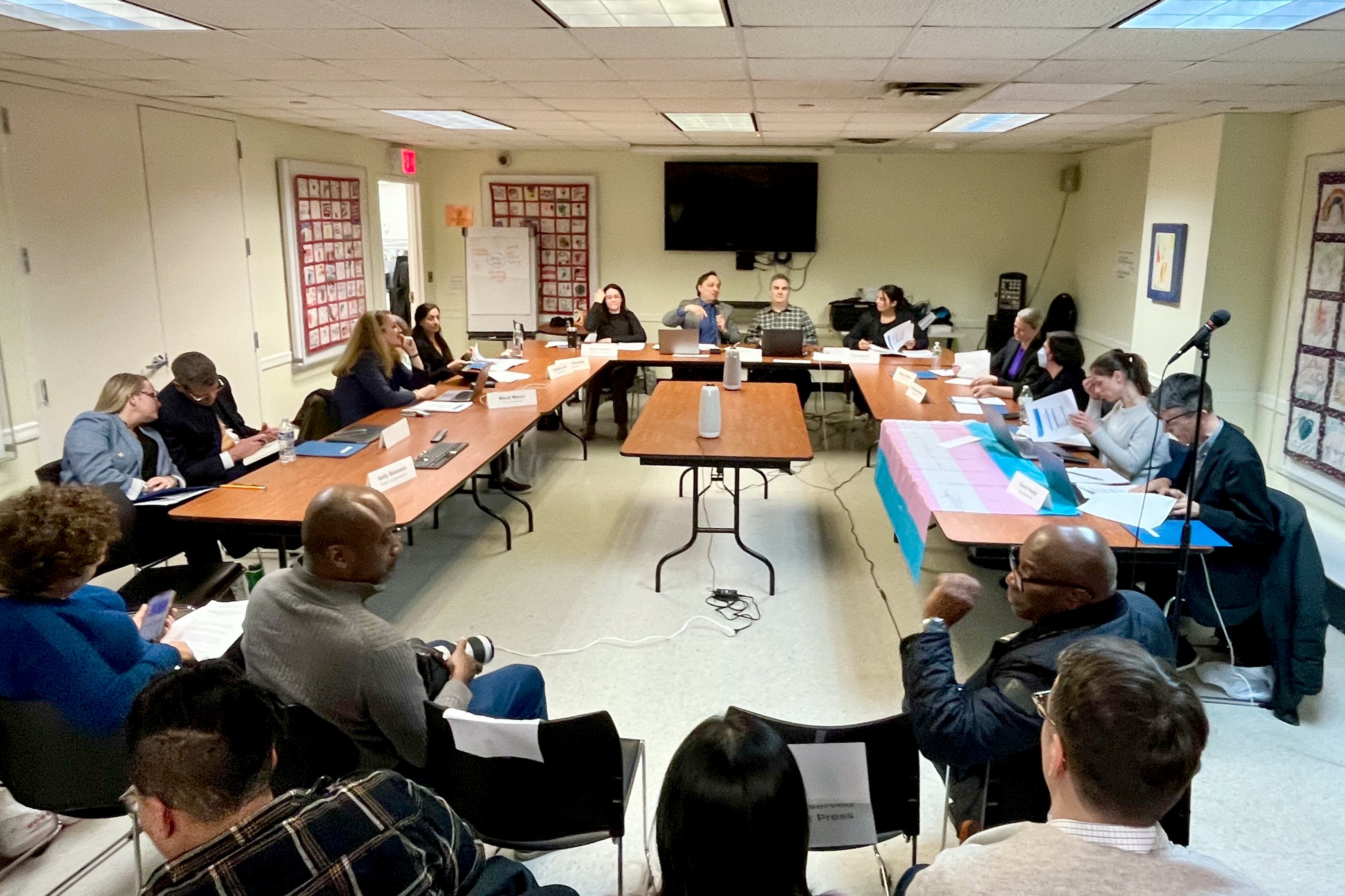 A group of people sit at tables in a rectangle in a room.