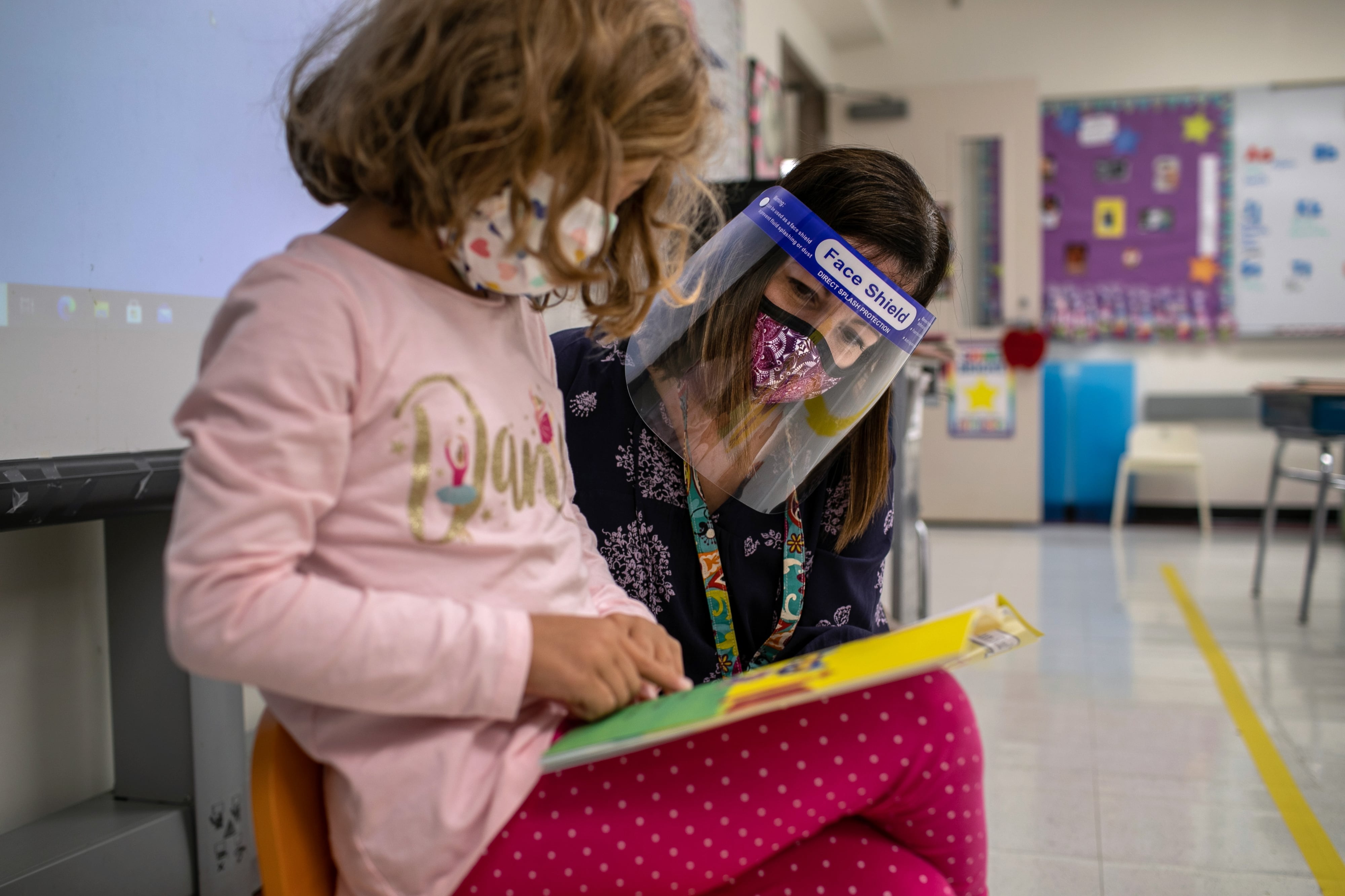 A young girl wearing a pink shirt and protective mask reads a book while her teacher, a woman wearing a face shield and mask, looks over her shoulder in a classroom.