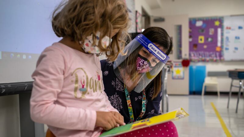 A young girl wearing a pink shirt and protective mask reads a book while her teacher, a woman wearing a face shield and mask, looks over her shoulder in a classroom.