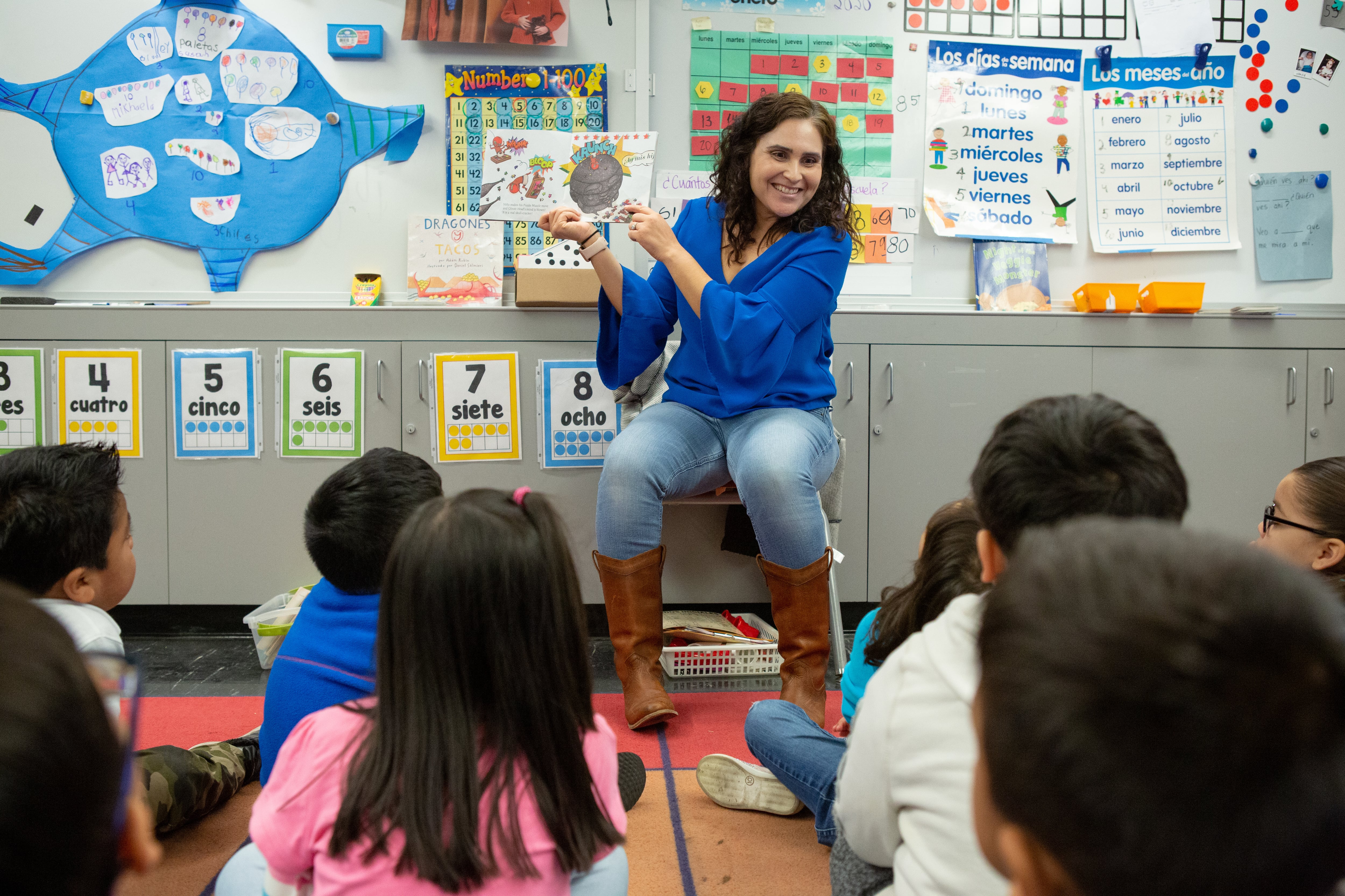 A teacher sits on a chair in front of her young students, showing them pictures in a book she’s holding.