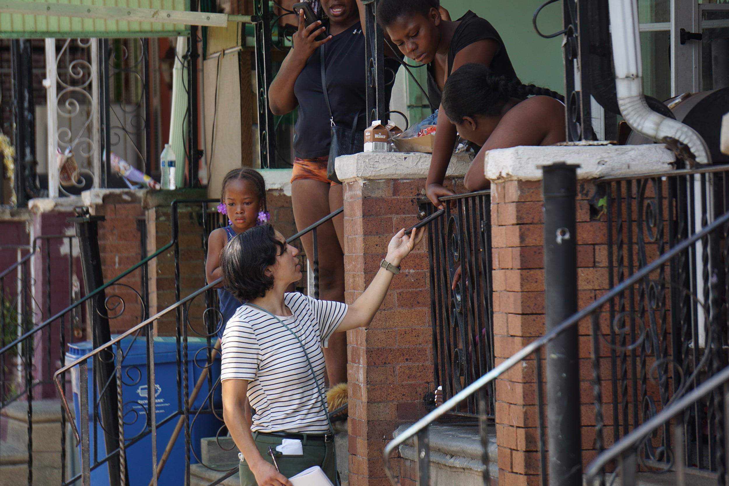 A photograph of a white women reaching out with her phone while speaking with four Black residents standing on a brick patio.
