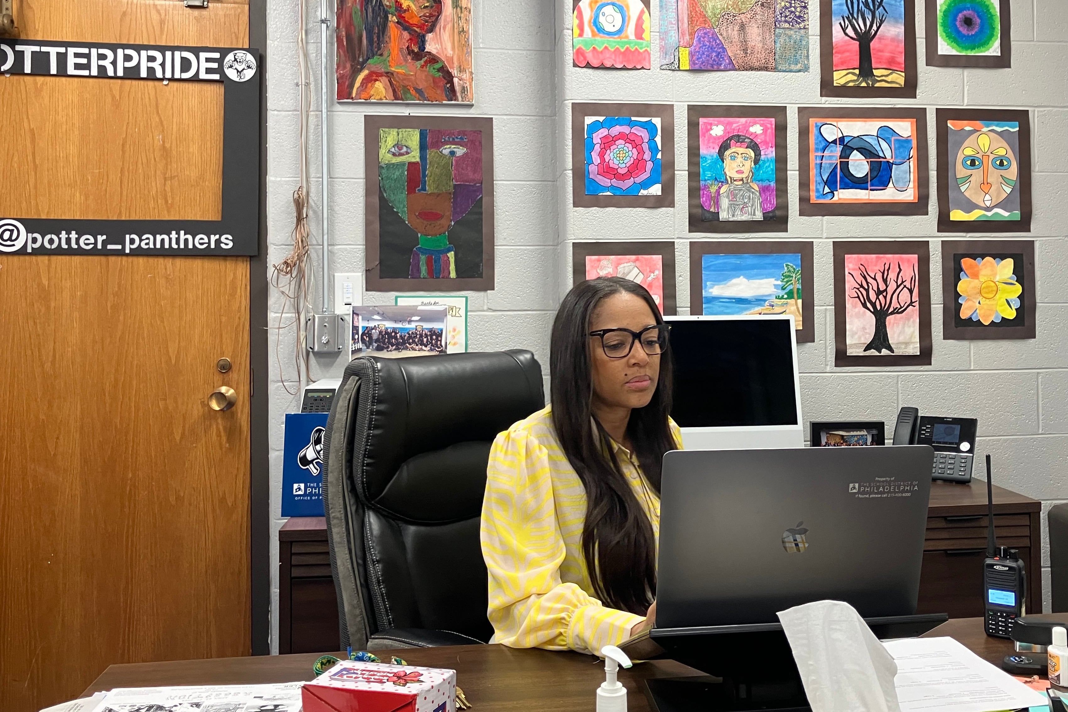 A Black woman with long, dark hair and wearing a yellow blouse sits behind a computer at a desk in a colorfully decorated office.