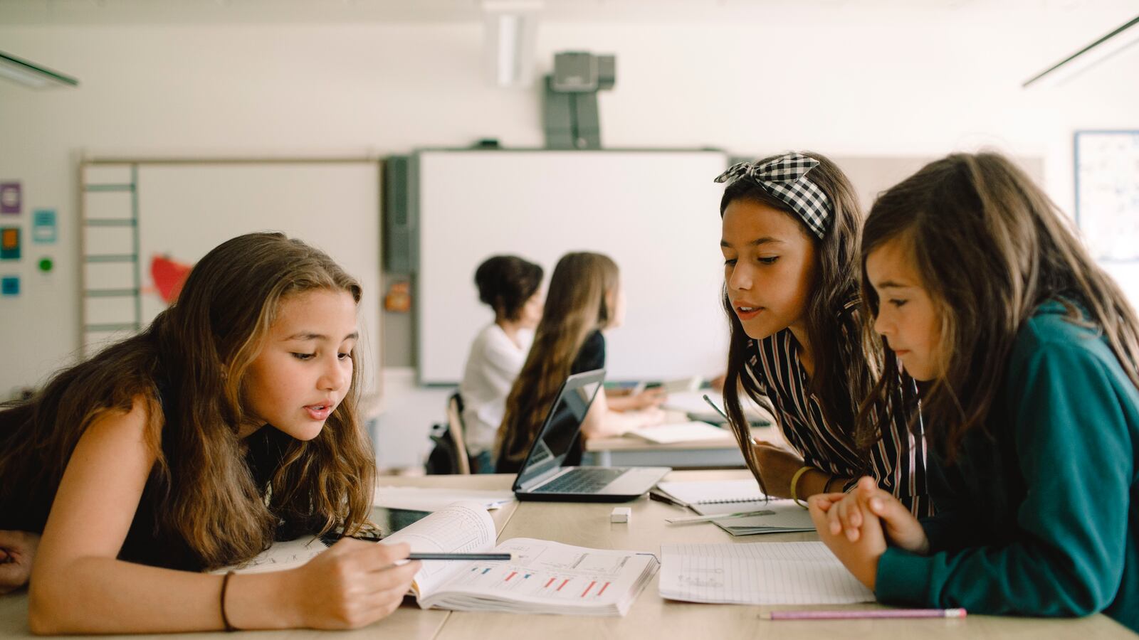 Three girls sit at a shared table in a classroom looking over an assignment in a book.