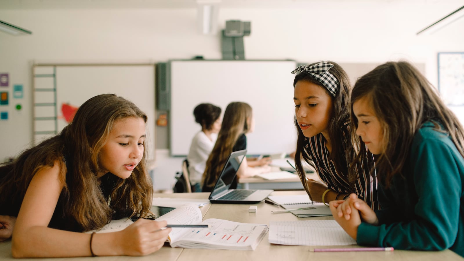 Three girls sit at a shared table in a classroom looking over an assignment in a book.