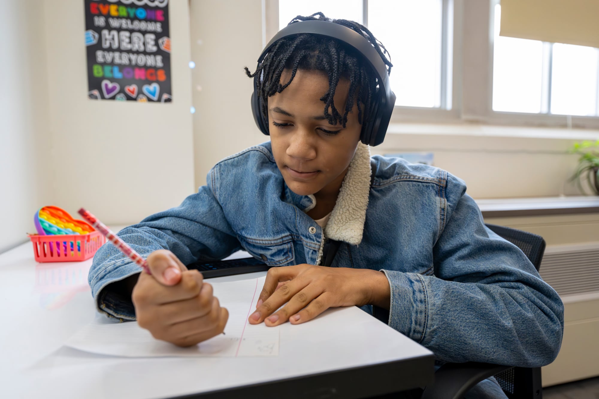 A boy wearing a jean jacket and headphones holds a pencil and writes