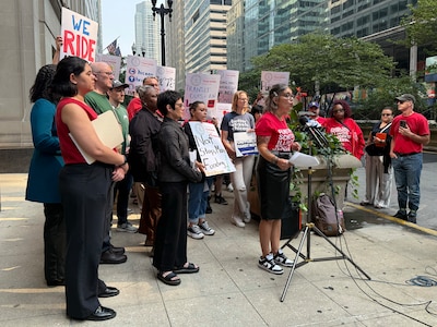 A photograph of a large group of people standing outside, some holding signs, while one person speaks from a microphone.