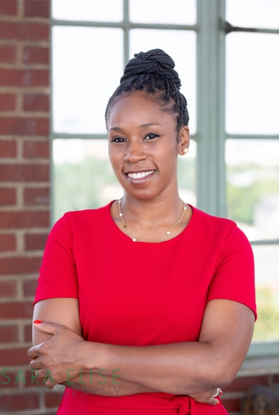 A Black woman with her hair tied back and wearing a red dress poses for a portrait in front of a brick wall and a glass window.