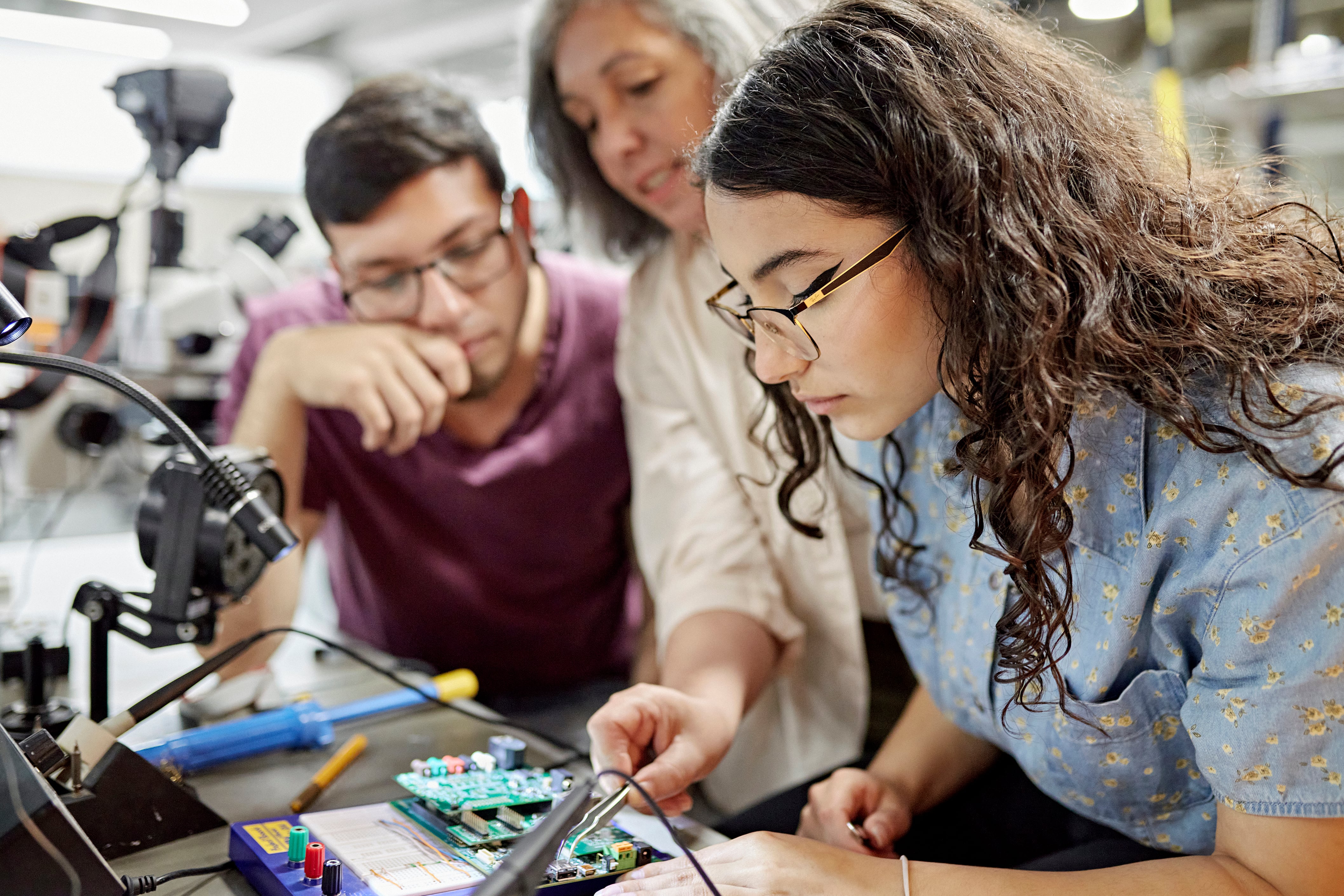 A woman in a jean shirt works on a mechanical device with switches while two others watch on.