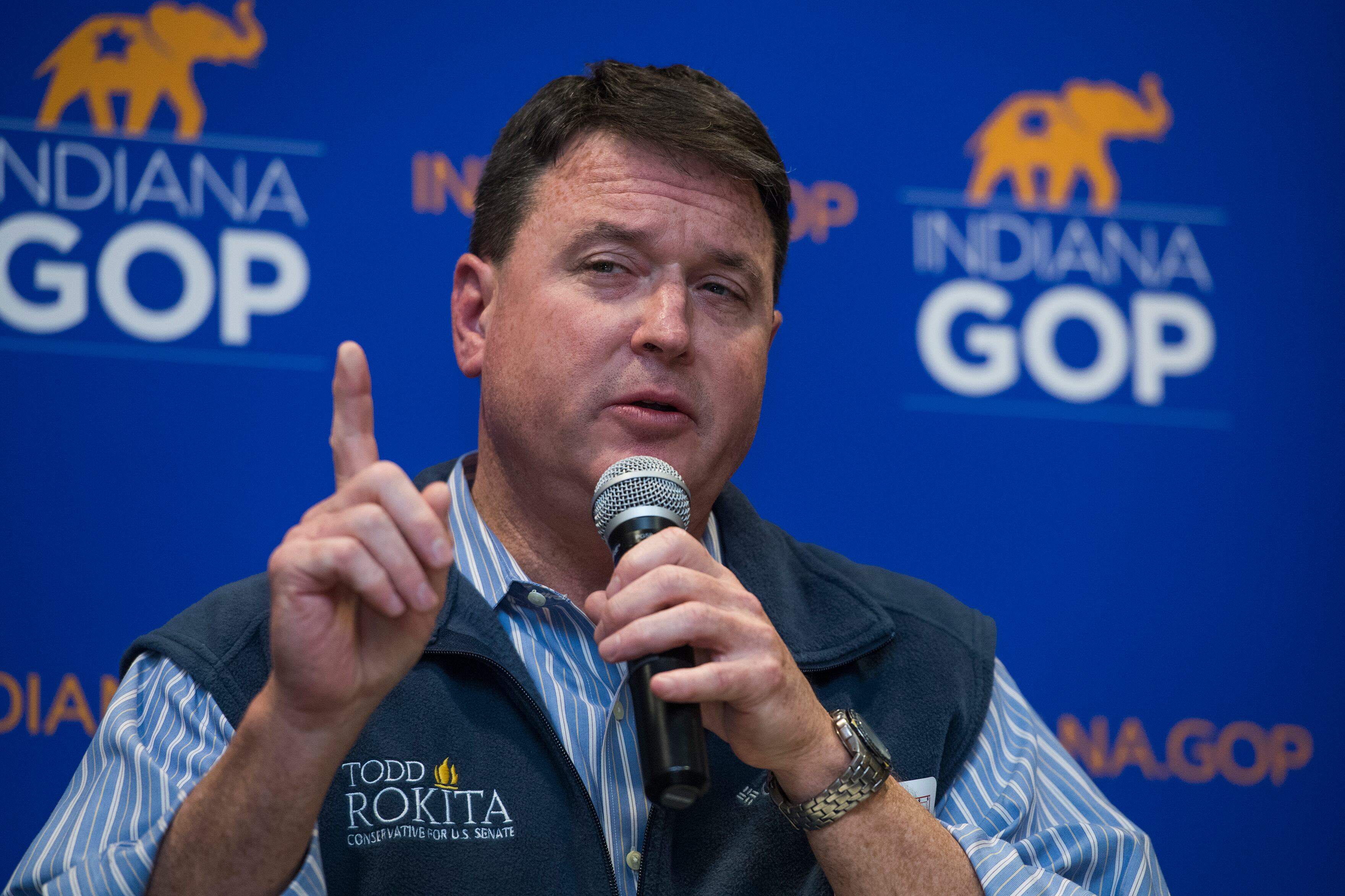 A man with short dark hair and wearing a dark vest over a blue and white stripe shirt holds a microphone and points with his hand. There is a blue background with small elephants and logos that read "Indiana GOP."