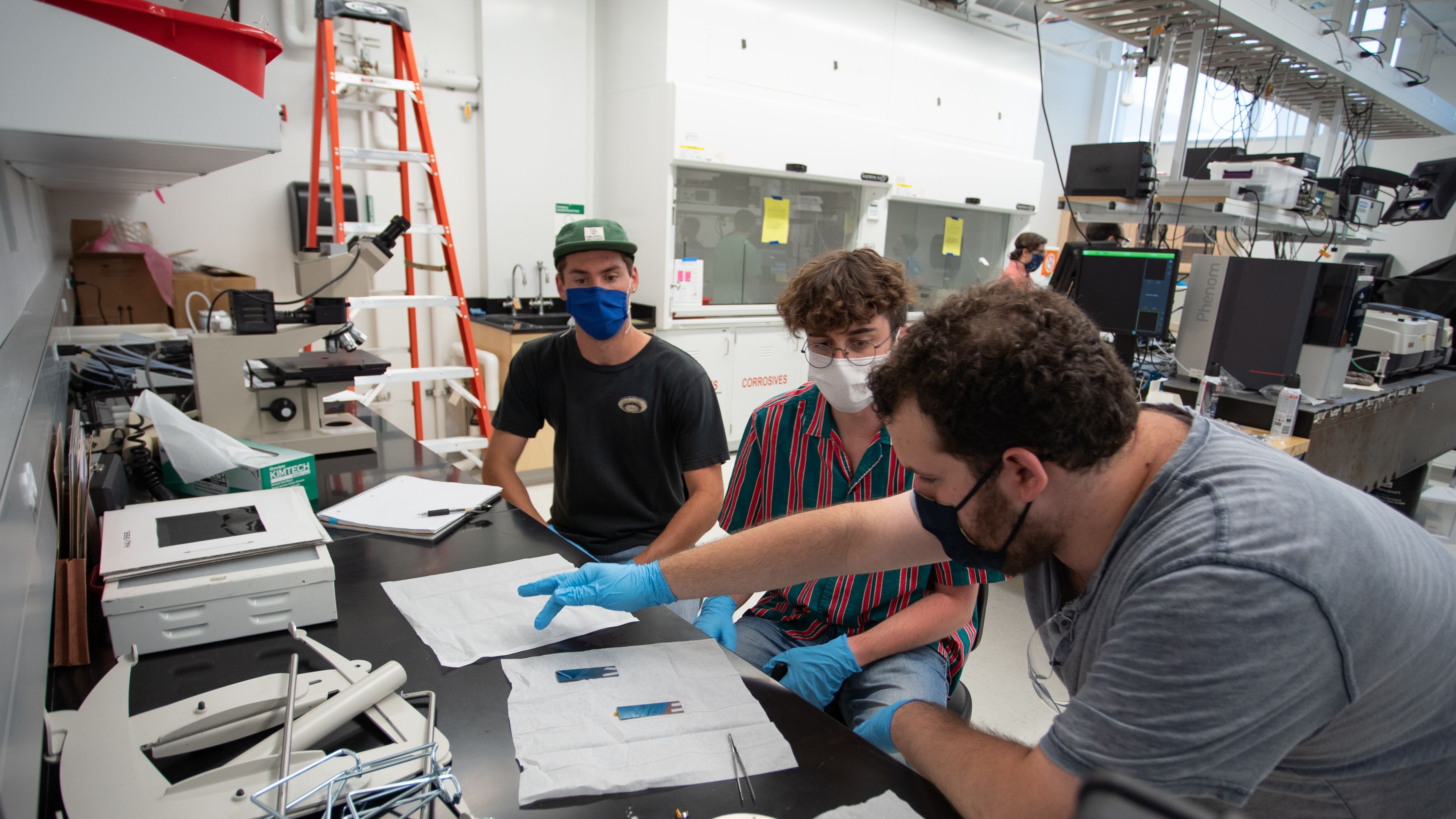 Three students wearing blue gloves gather around a work table in a lab class at the Colorado School of Mines. One student points at a paper.