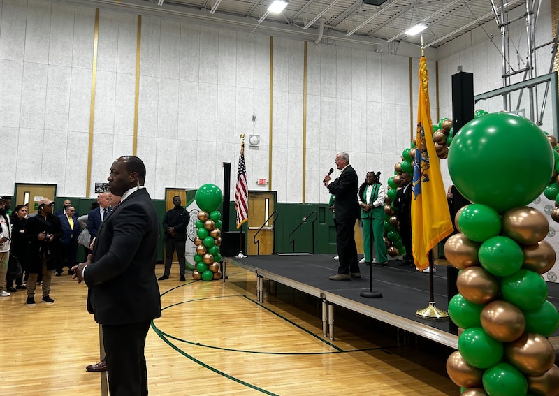 A white man in a suit speaks from a podium in a school with balloons on both sides of the stage.