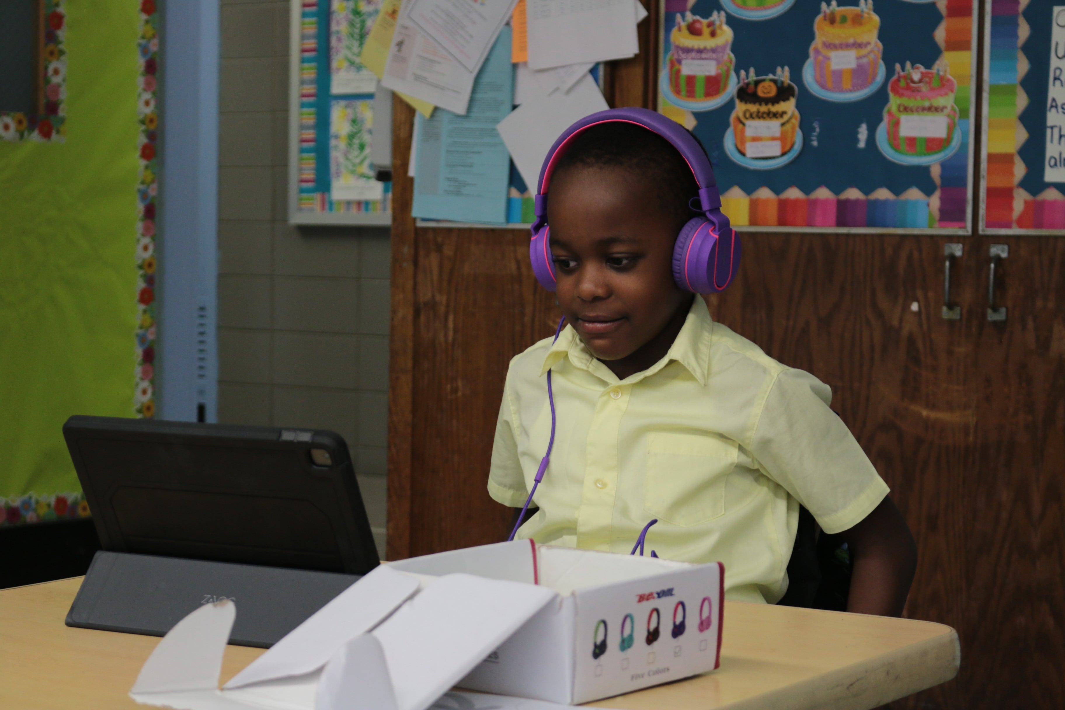 A young student wears headphones while sitting at a desk and working on a laptop inside of a classroom.