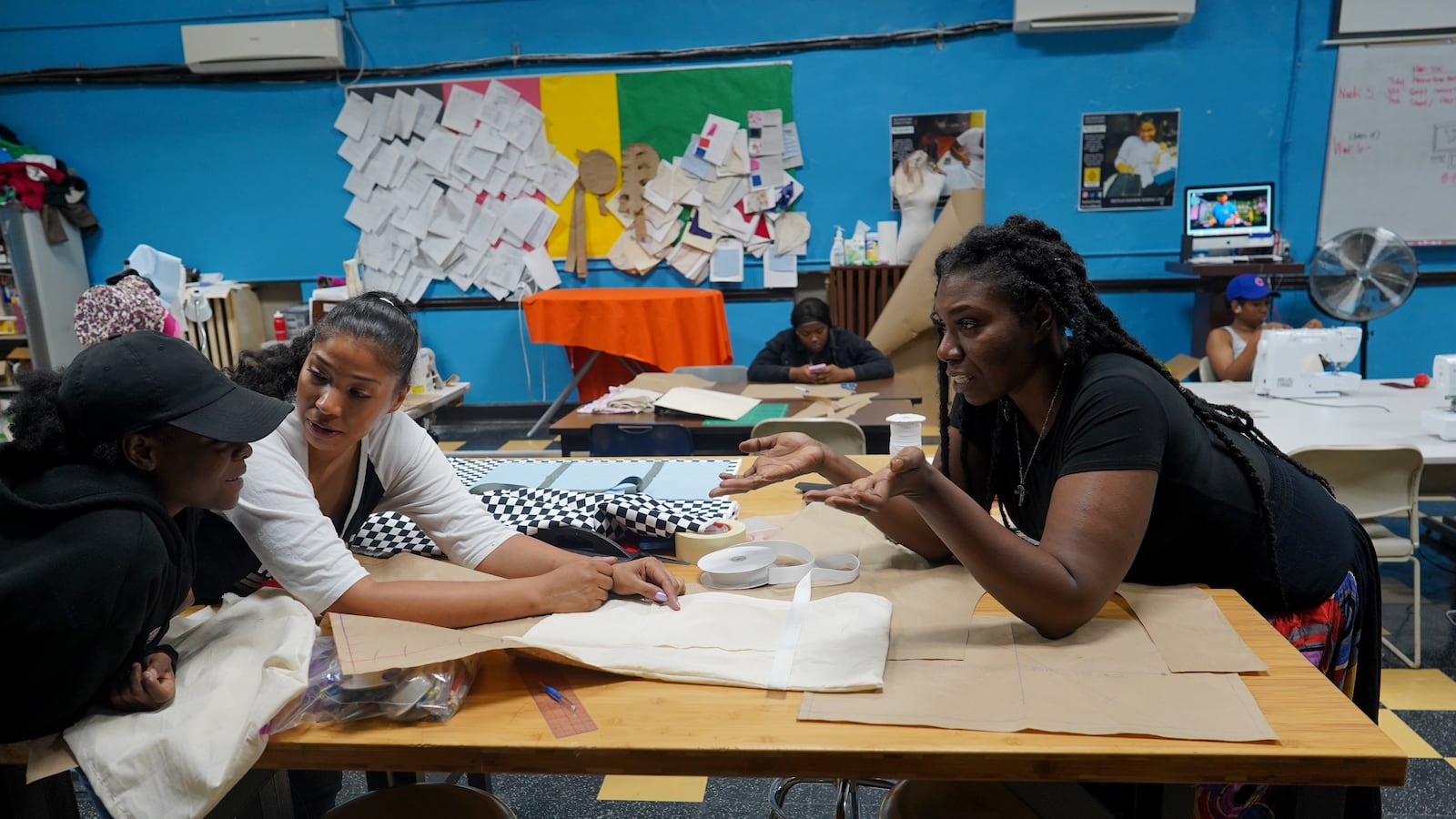 Two faculty members help a student inside of a school sewing lab, as other students work in the background