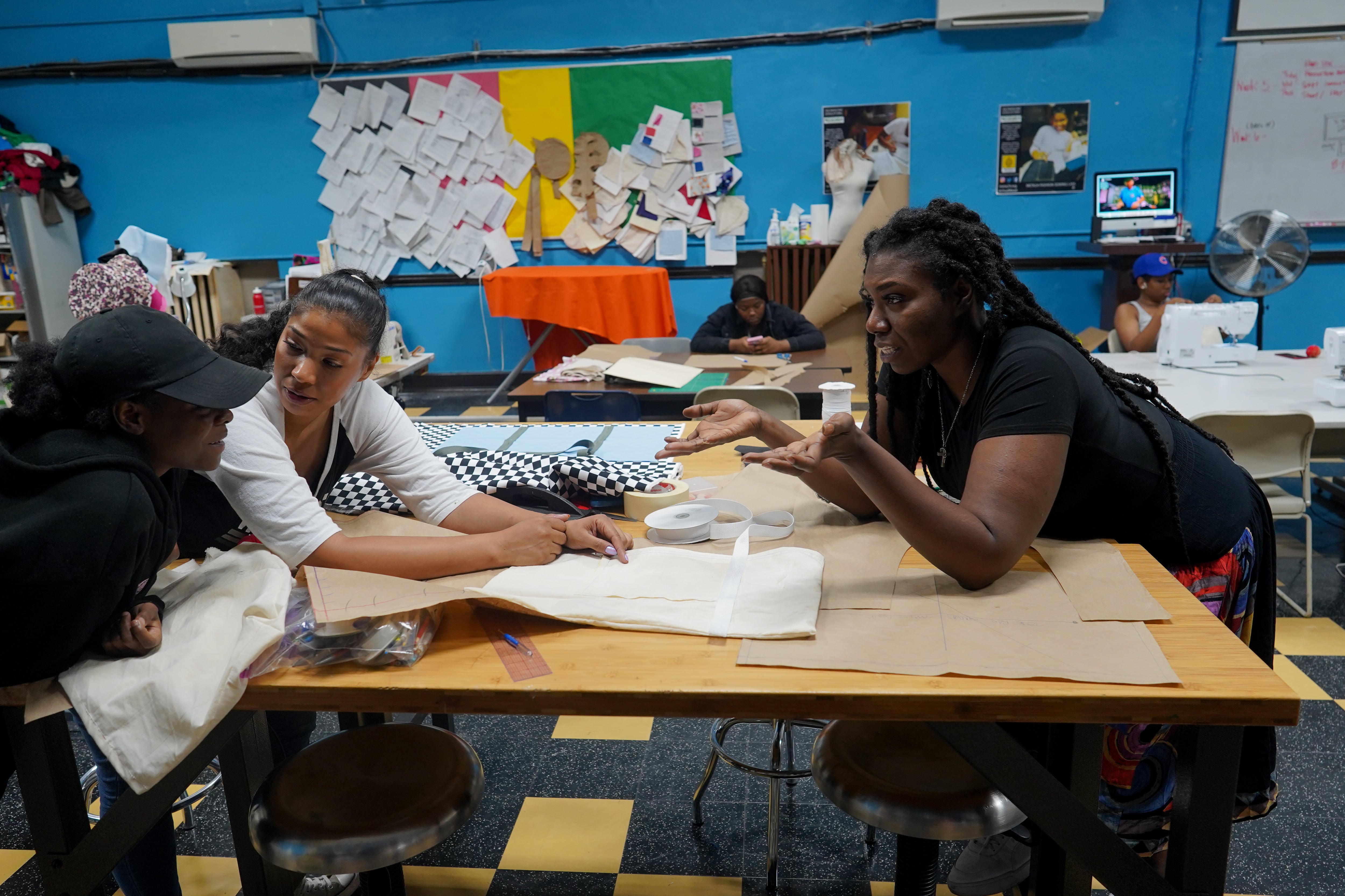 Two faculty members help a student inside of a school sewing lab, as other students work in the background