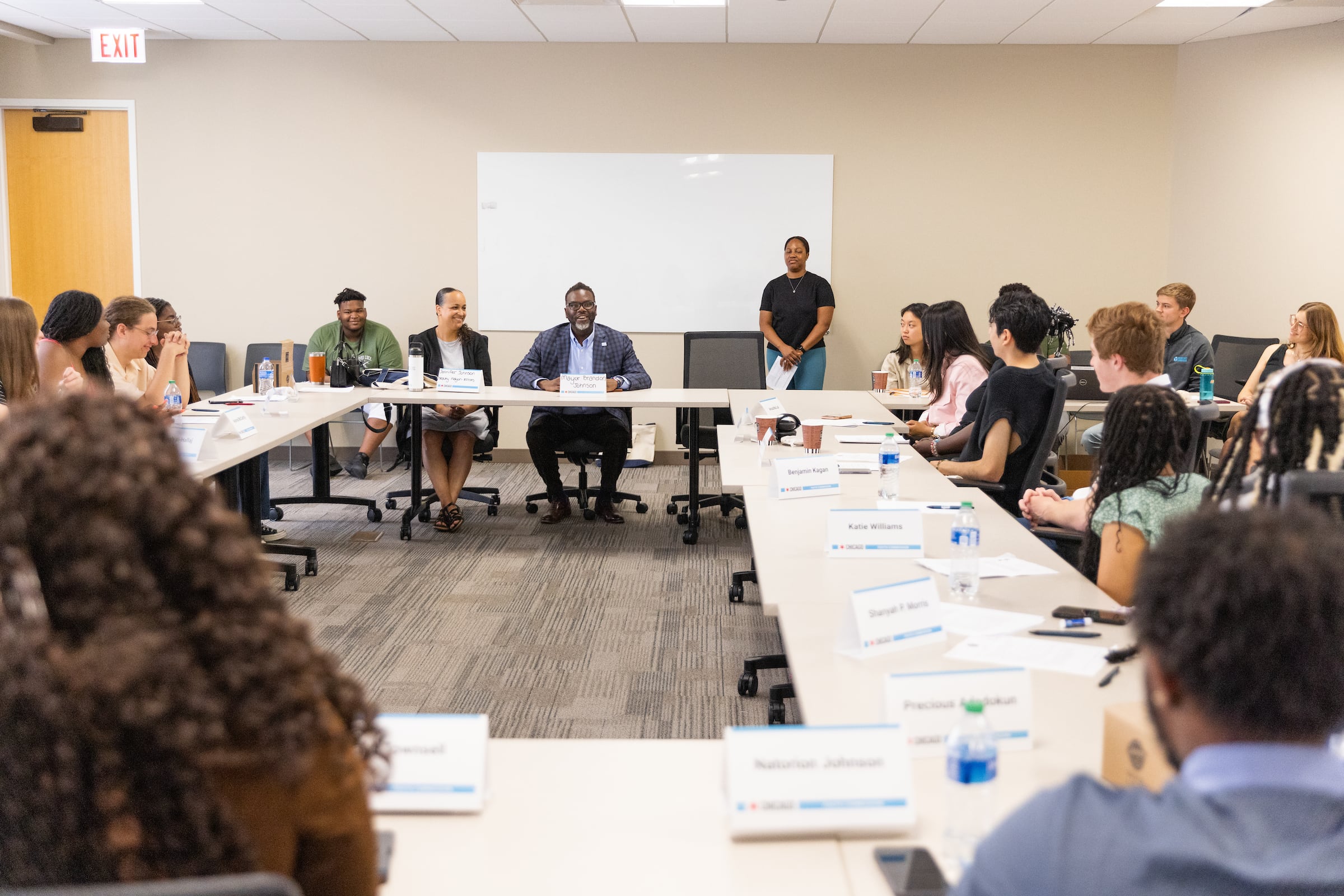 A group of teens and city officials sit around white conference room tables with papers and name plates in front of them.