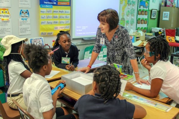 A woman in a black patterned dress talks to students at a table.