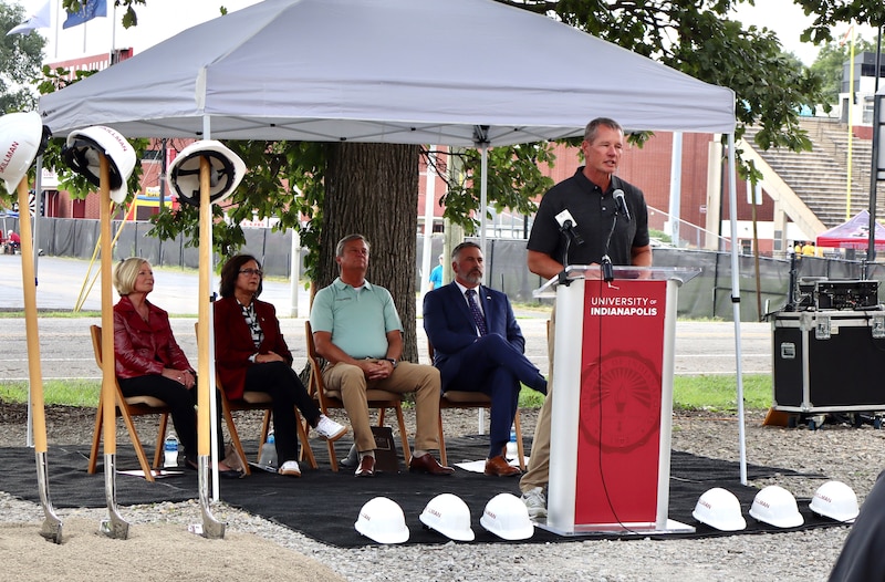 A man in a dark shirt speaks from a red-and-white podium in front of other people seated under a white tent.