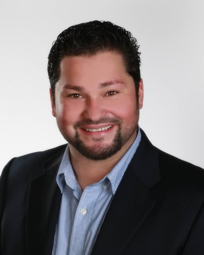 Stephen Varela smiles for a formal portrait. The photo is a headshot of a late 30s Hispanic man in a dark blazer and open collared blue dress shirt. He has dark hair and a closely cropped beard.