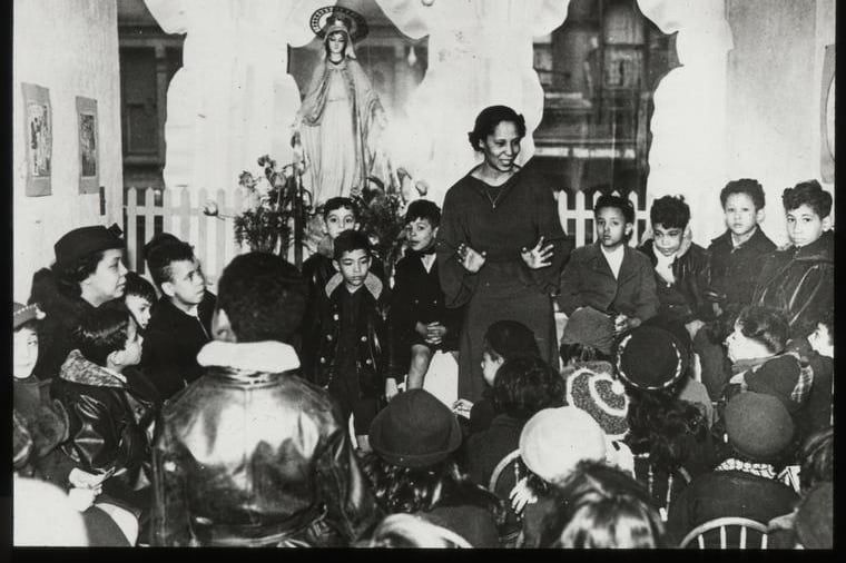 A black and white historic photo shows a woman talking to a group of children.