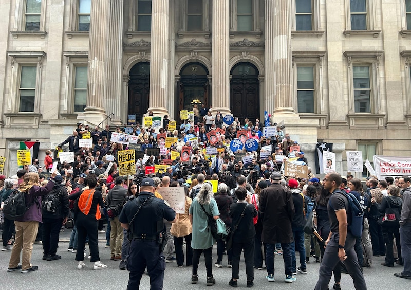 A crowd of people, some holding signs, stand on the steps of a building with pillars.