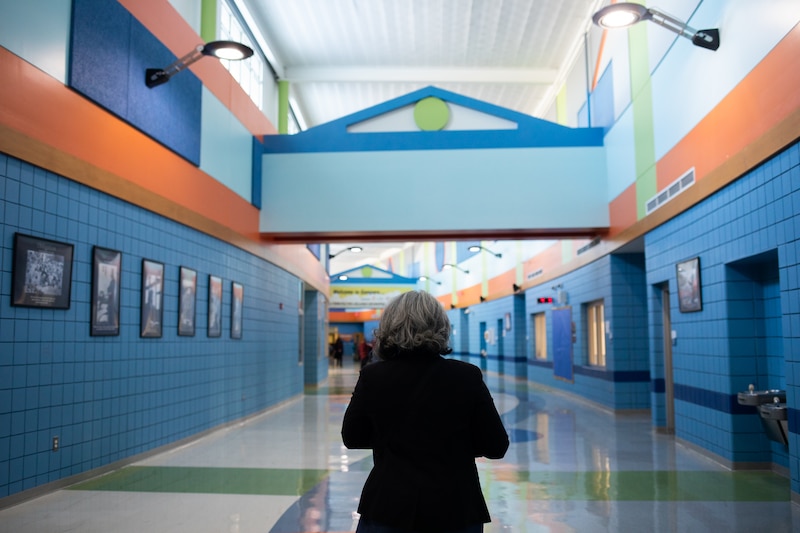 A woman with short hair and wearing a dark suit stands in the middle of a school hallway.