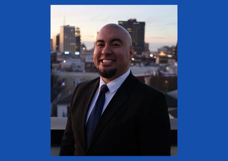 A man in a suit stands against the backdrop of a city skyline.