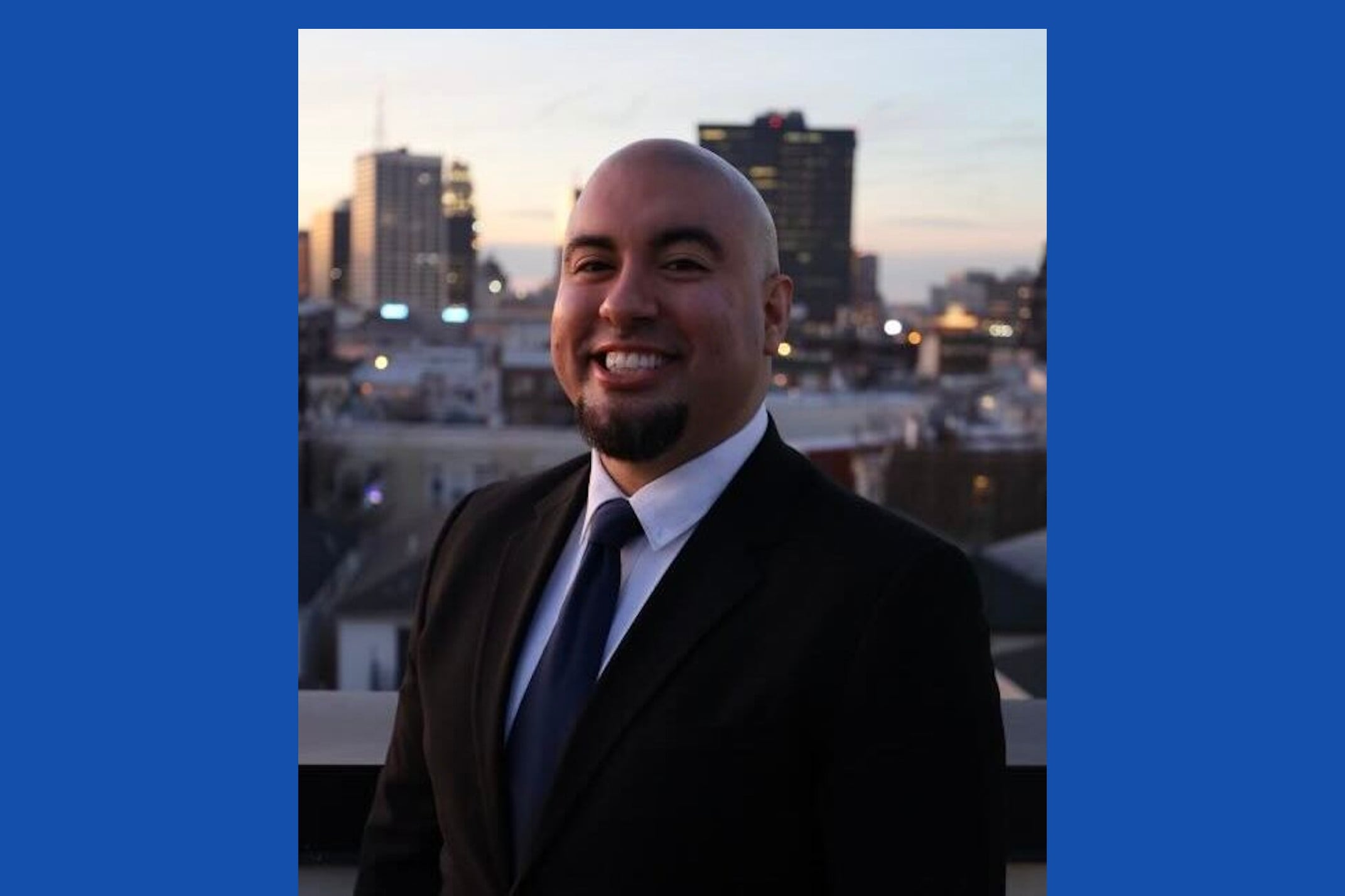 A man in a suit stands against the backdrop of a city skyline.