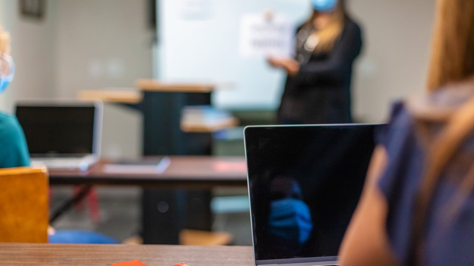 A female teacher wearing a face mask and holding up signs in front of the class in a classroom setting.