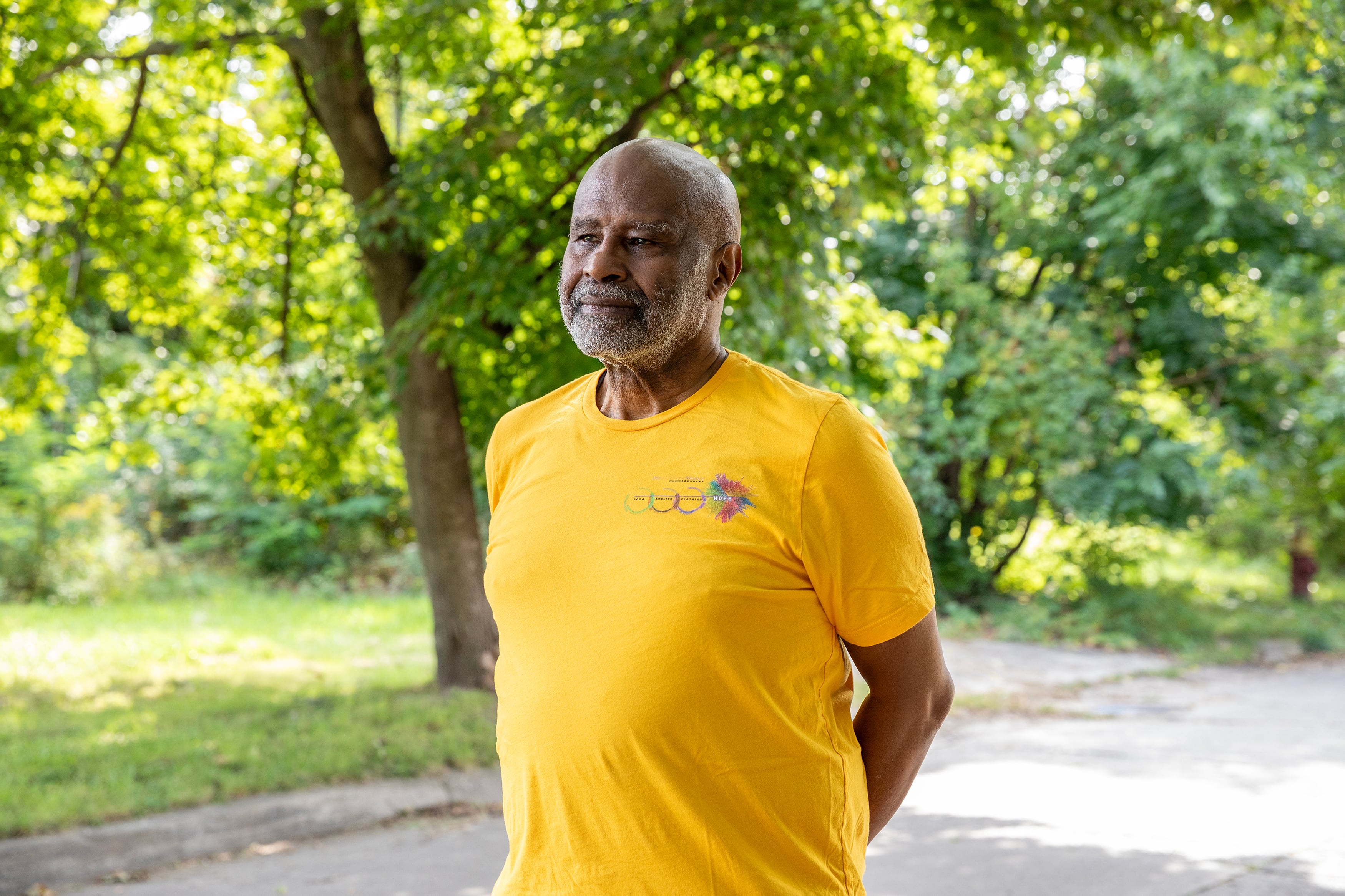 A man wearing a yellow shirt stands poses for a portrait with green trees in the background.