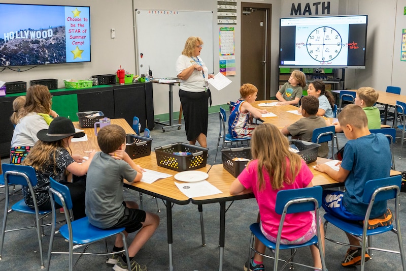 A teacher stands in between young students at school desks in a classroom.