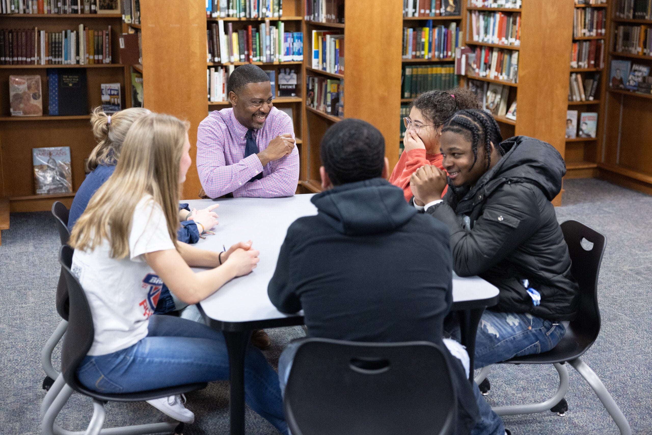 A Black male teacher smiles while sitting at a table and looking at a group of five high school students of different races.