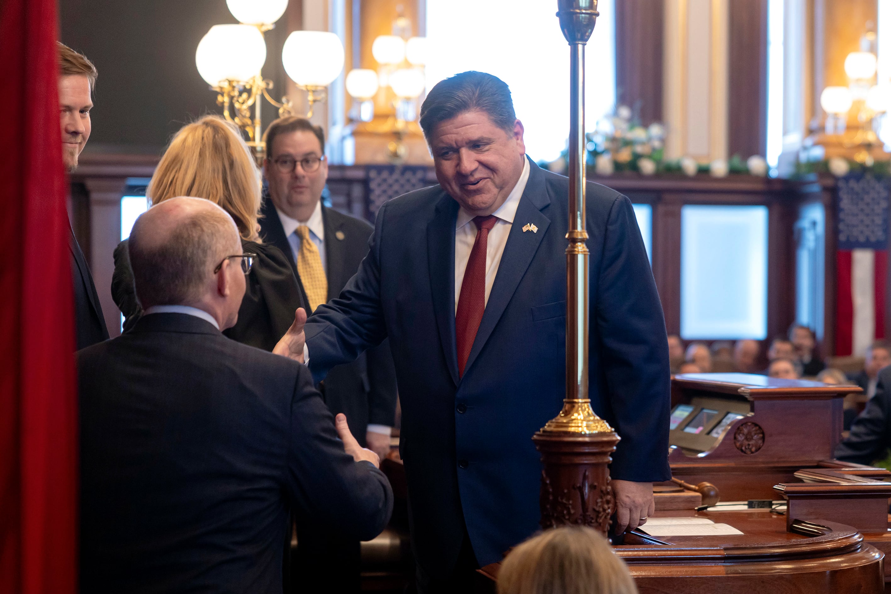 A man in a blue suit and red tie greets another man in a suit in a statehouse room.