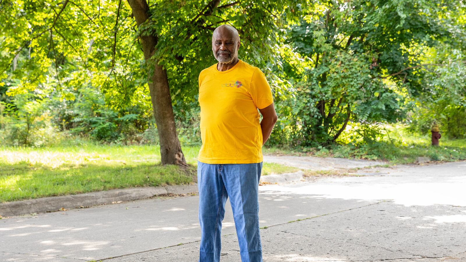 An adult man wearing a yellow shirt and blue jeans poses for a portrait in the middle of a street with green trees and grass in the background.