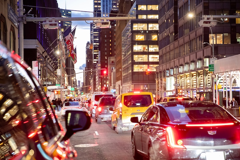 Cars are stopped in traffic on a city street with buildings on each side of the photo. Red break lights fill the bottom half of the photo with a sliver of sky in the background.