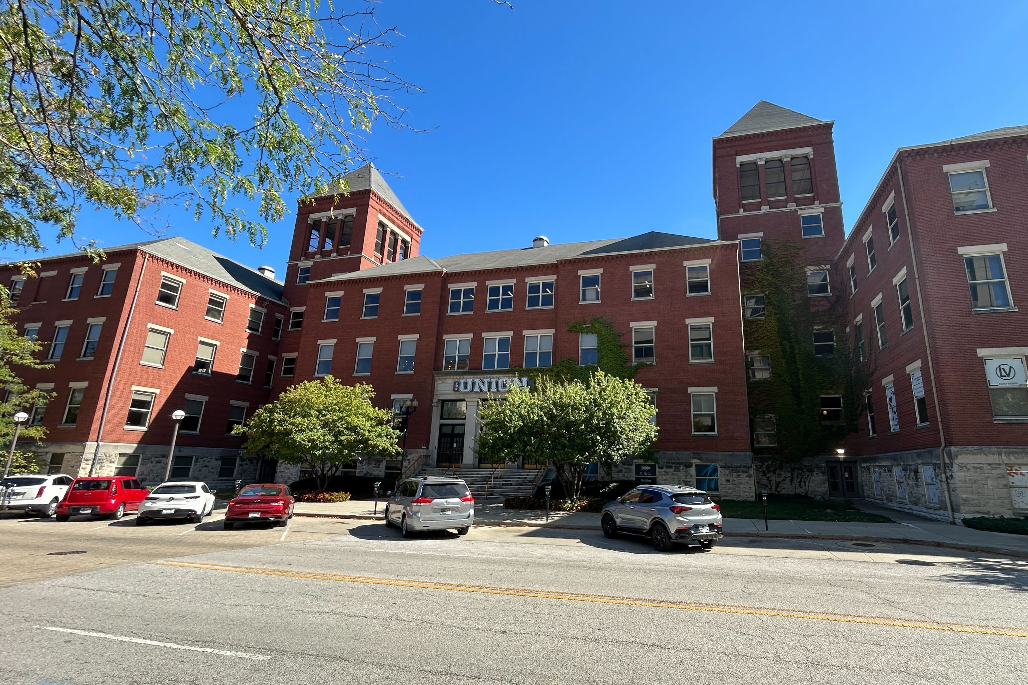 A photograph of the exterior of a large red brick building with green trees and parked cars out front with a blue sky in the background.